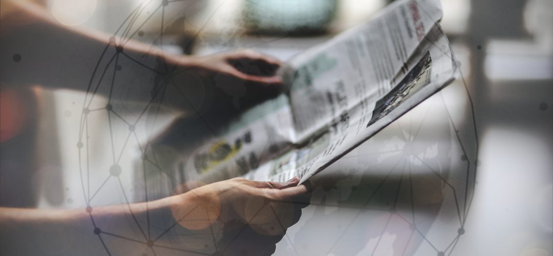 Duas mãos segurando um jornal impresso aberto. Com uma marca de transparência, um globo aparece por cima da imagem de forma sutil