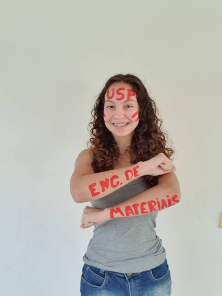 Menina com o os braços e rosto pintados de vermelho, com o nome da universidade e do seu curso.