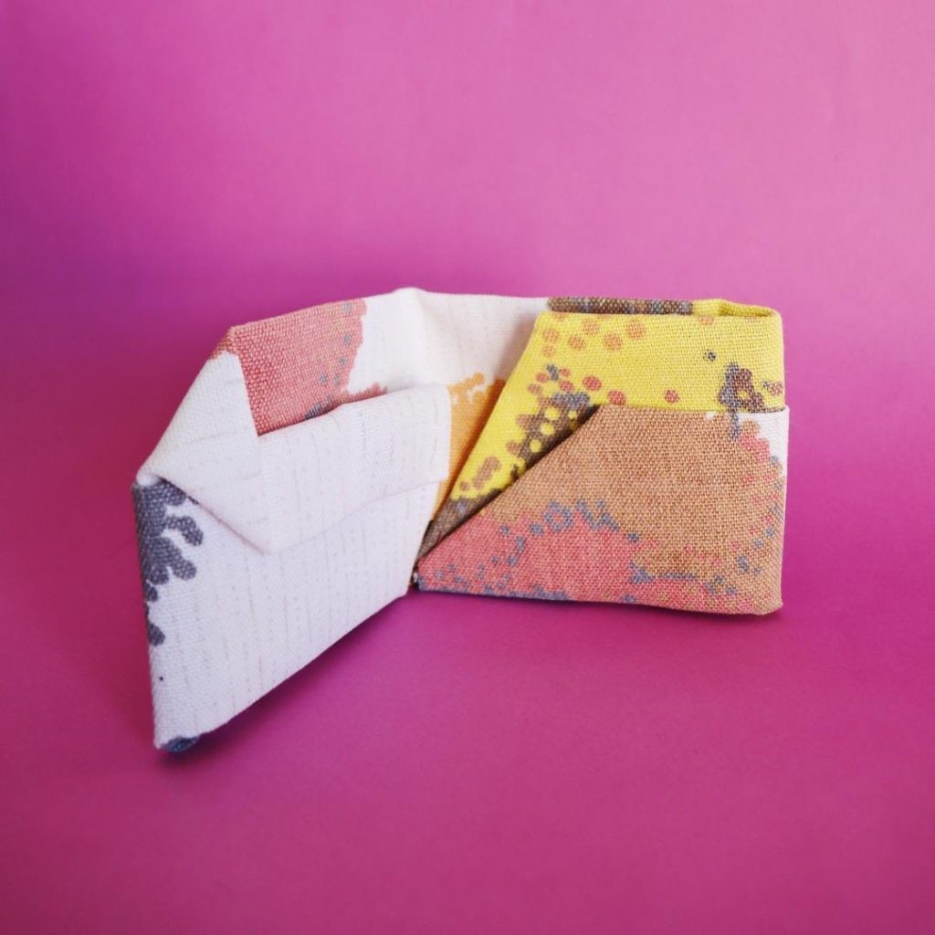 Foto quadrada de fundo rosa. No centro da imagem há uma carteira feita de tecido estampado