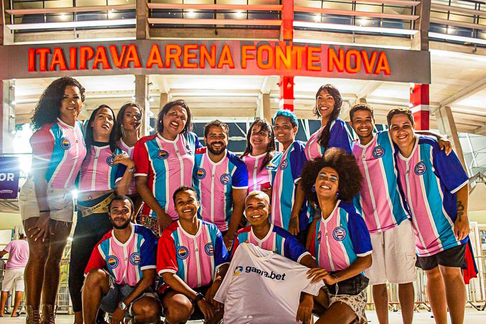 Grupo de torcedores vestidos com camiseta do time Bahia estampadas com a bandeira trans, nas cores branco, azul e rosa