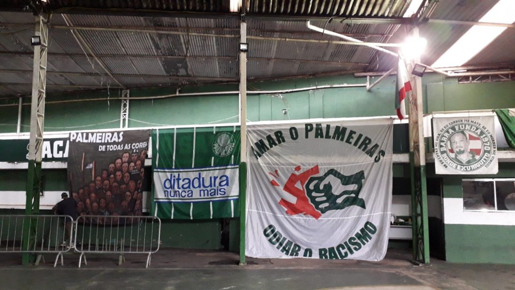 Galpão com três bandeiras estendidas. A primeira, à esquerda, é uma releitura da obra Operários, de Tarsila do Amaral, com a frase "Palmeiras de todas as cores". Ao centro, outra faixa listrada de verde e branco com a frase "ditadura nunca mais" e o brasão do palmeira no canto superior direito. À direita, uma bandeira branca escrito "amar o palmeiras, odiar o racismo", com um desenho ao centro.